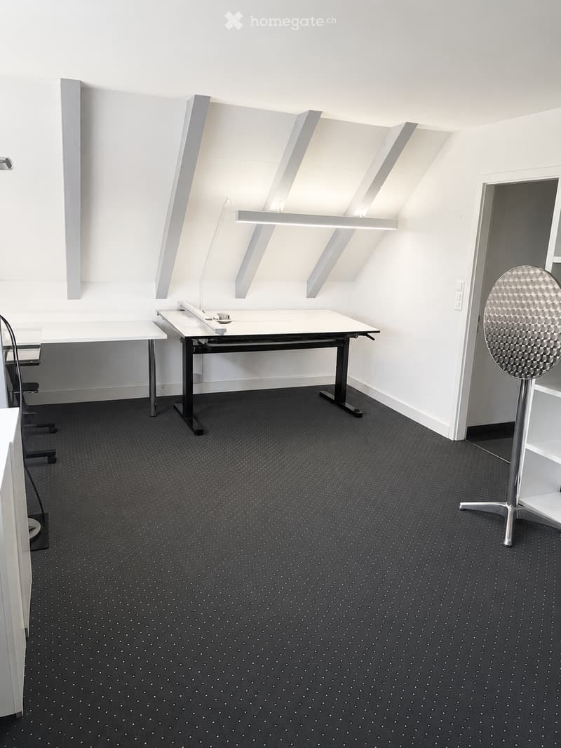 Modern, minimalist room with white walls, grey carpet, and built-in shelving. Contains a white table, chair, and a round metallic stool.