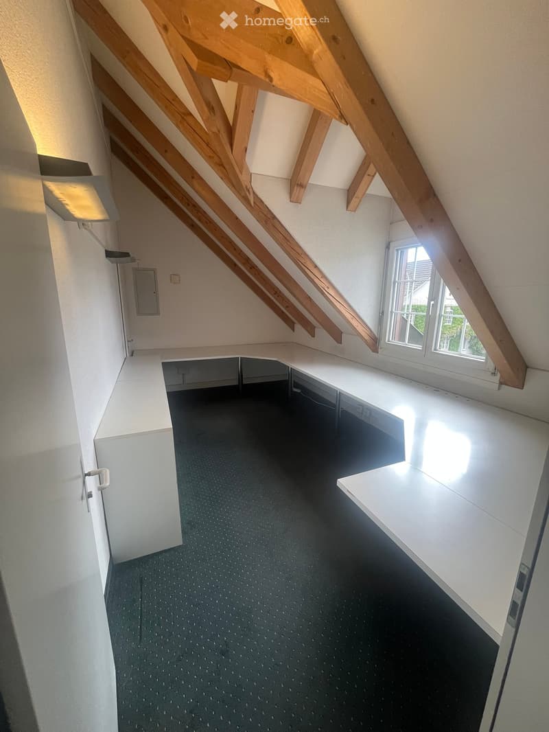 An attic space with exposed wooden beams, a large window, and built-in white shelving units along the walls. The floor is covered with a dark carpet, and the walls are painted white.