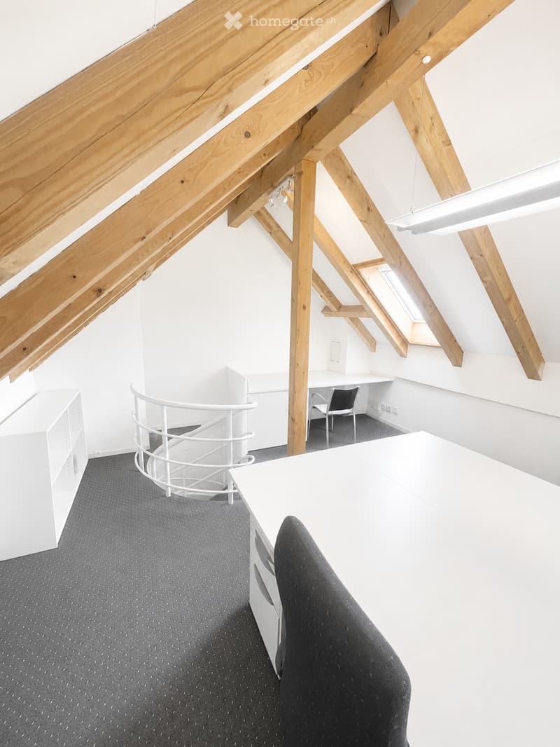 Modern office space with exposed wooden beams, white walls, grey carpet, and contemporary furniture including a desk, chair, and shelving unit.