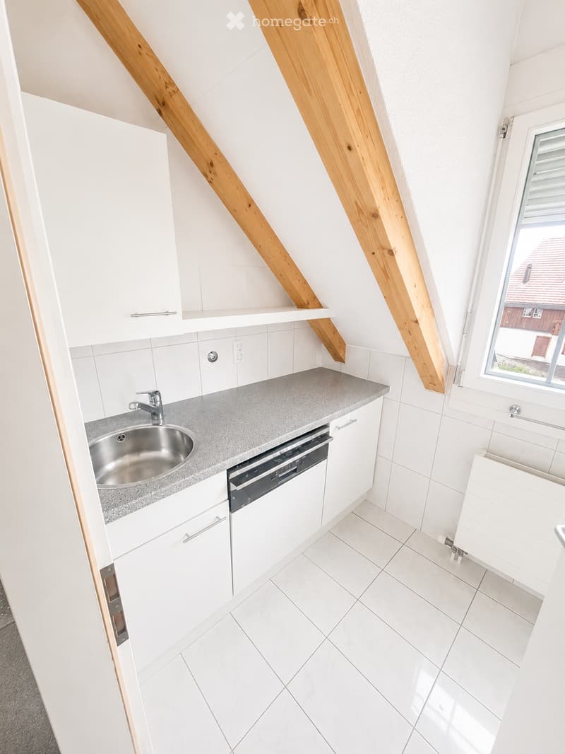 Modern kitchen with white cabinets, grey countertop, stainless steel sink, and built-in dishwasher. The room features white tiled walls and floor, with wooden beams on the ceiling and a window offering a view of the exterior.