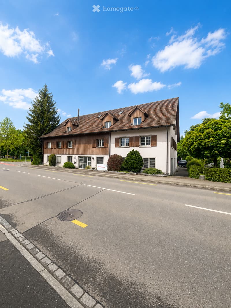 Two-story house with a brown tiled roof, white walls, and multiple windows with shutters. The house is surrounded by greenery and is located on a quiet street.