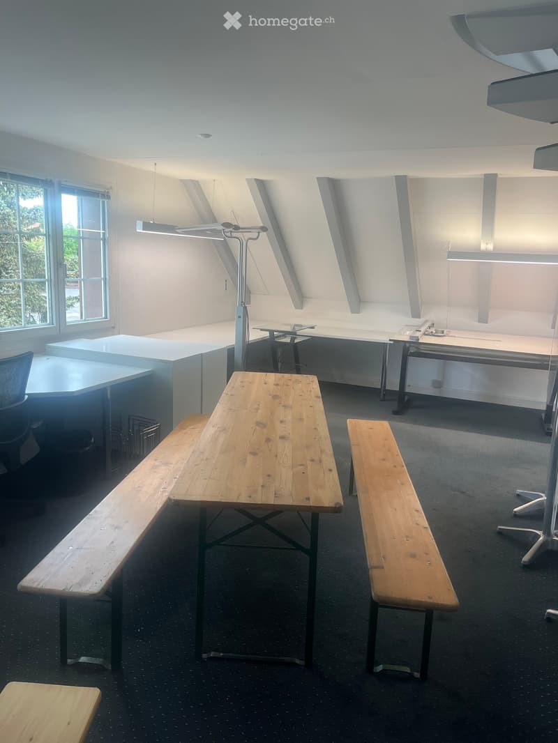 Bright, modern room with large windows, wooden benches, tables, and a desk setup.