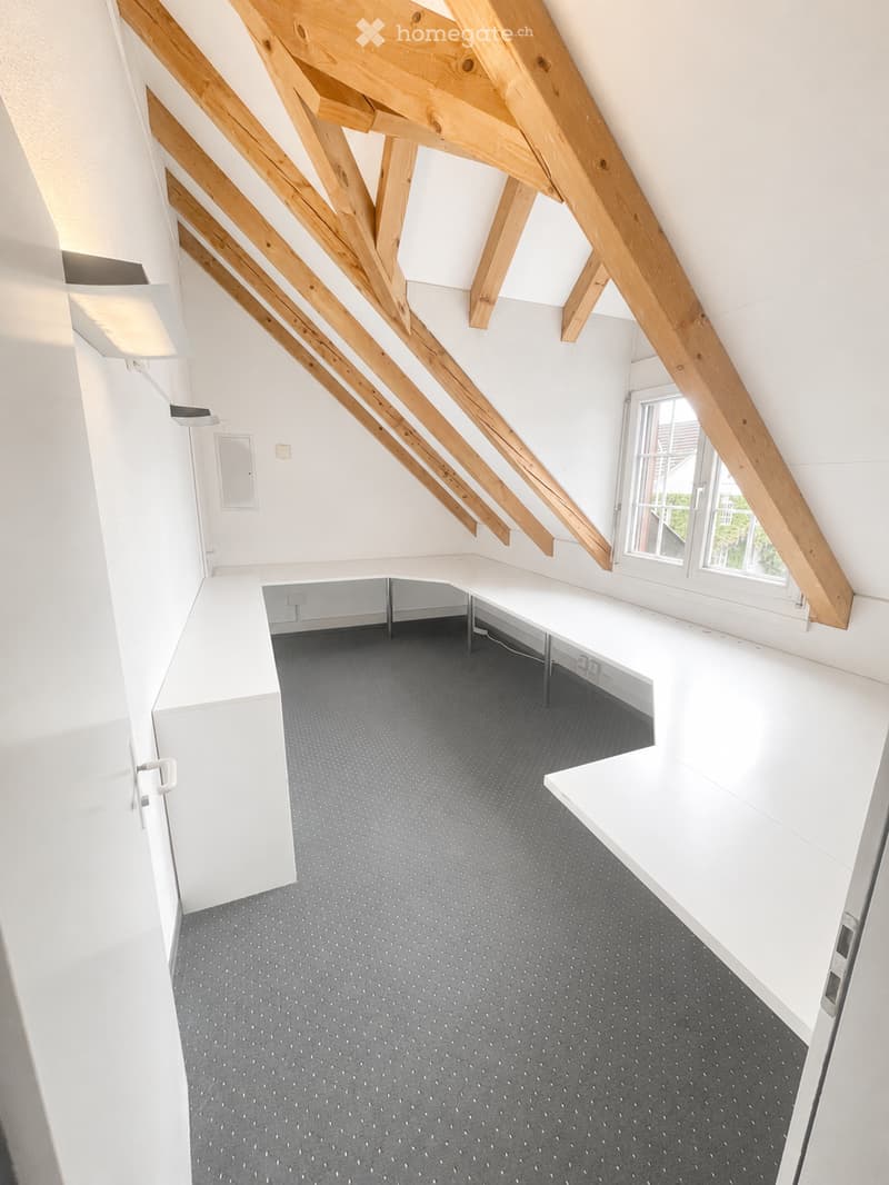 An empty attic space with exposed wooden beams, grey carpeted floor, white walls, and a large window letting in natural light.