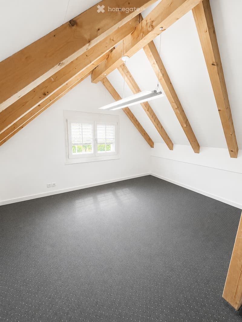 Empty room with exposed wooden beams, grey carpeted floor, white walls, and a single window with blinds.