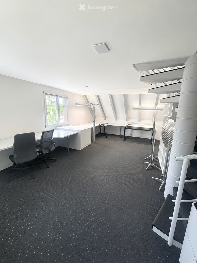 Modern office space with grey carpet, white walls, large windows, desks, chairs, and shelves.