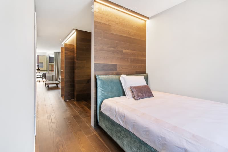 Bedroom with wooden floor, white walls, bed with white sheets and pillows, wooden wall paneling, and a small hallway leading to another room.