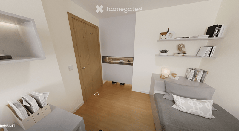 A modern, compact living space with a grey sofa, decorative pillows, built-in shelves holding books and decorative items, a small table with a lamp, and a small kitchen area visible through an open doorway.