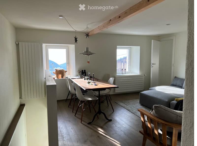 Furnished Room. A modern, open-concept living and dining area with light-colored walls, wooden flooring, and large windows. Features include a sofa, a dining table with chairs, and a hanging pendant light.