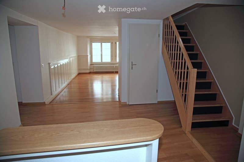 Bright hallway with wooden flooring, white walls, a staircase with wooden railings, and a small wooden bench in the foreground.