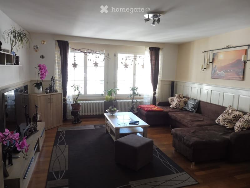 A modern living room with a dark brown sectional sofa, a glass coffee table, an ottoman, a wooden cabinet, and several decorative plants. The room features large windows with sheer curtains, a dark area rug, and wall-mounted candle holders.