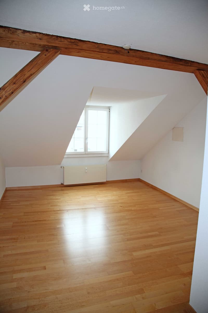 Empty attic room with wooden floors, white walls, exposed wooden beams, and a single window letting in natural light.