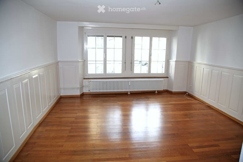 Empty room with wooden flooring, white walls, large windows, and a radiator below the windows.