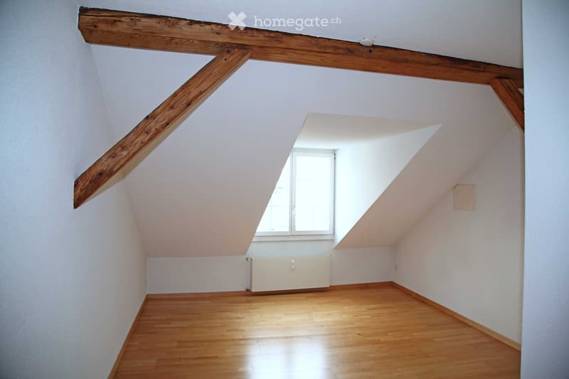 Empty attic room with wooden beams, wooden flooring, white walls, and a single window letting in natural light.