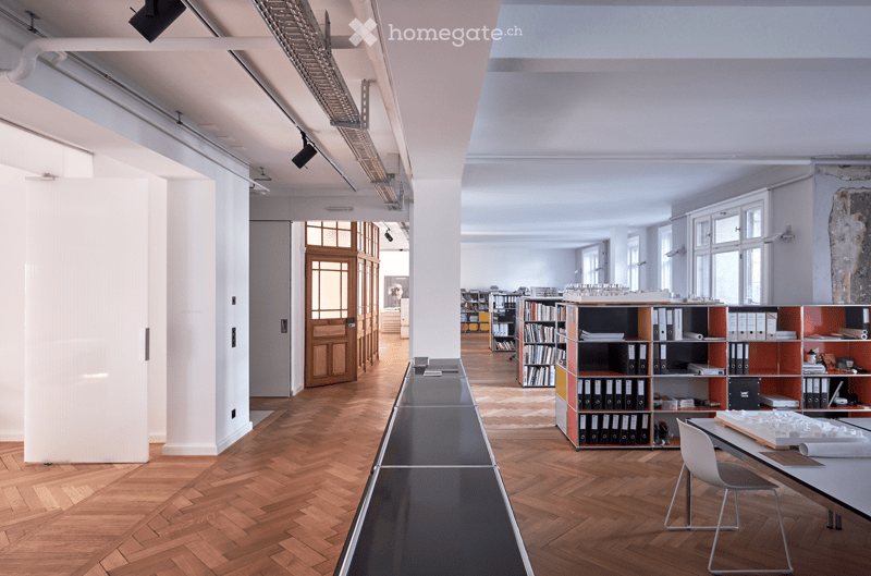 wooden parquet flooring, long rectangular counter, white walls, bookshelves, windows with wooden frames, desk with objects on top