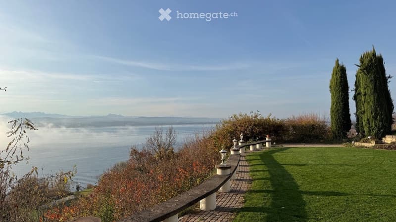 Garden / Outdoor Area. A serene garden area overlooking a lake, featuring a curved stone bench, neatly trimmed grass, and tall cypress trees.