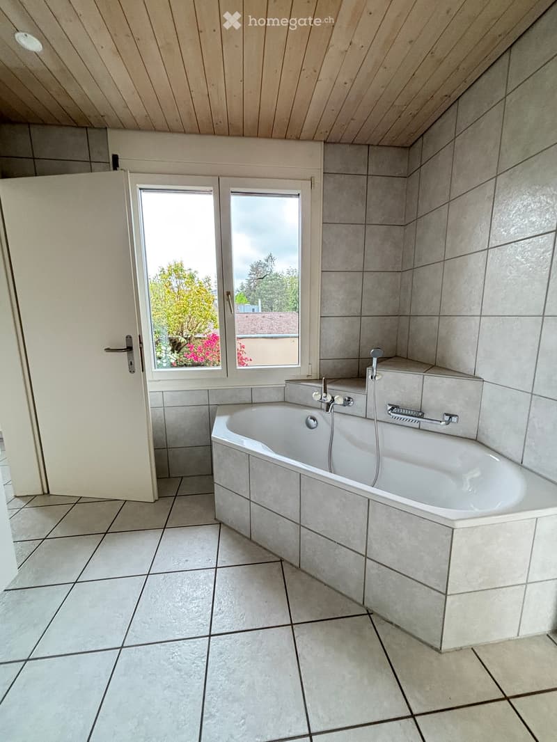 Modern bathroom with a bathtub, tiled walls and floor, and a window offering a view of greenery outside.