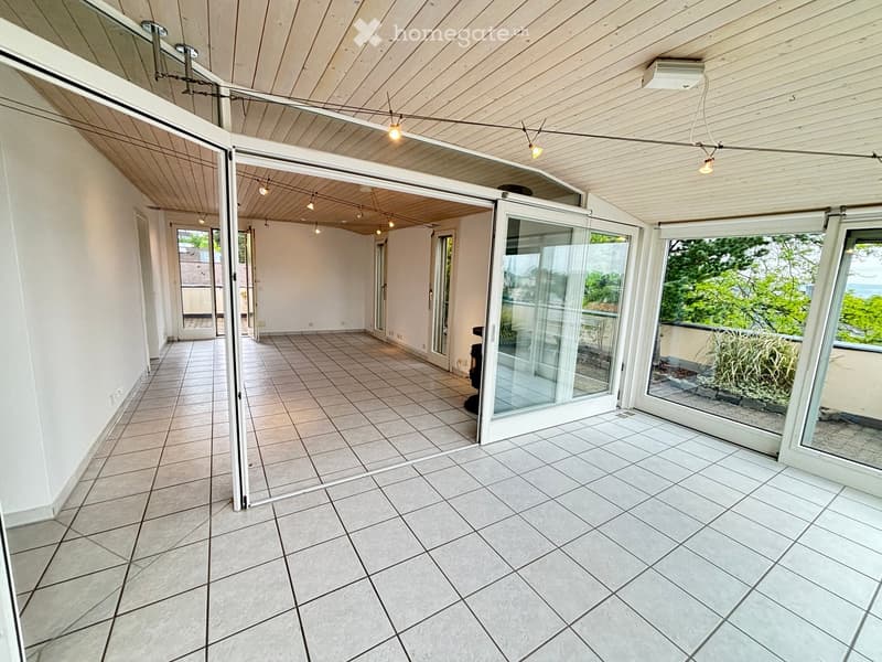 Bright, spacious balcony with large sliding glass doors, tiled flooring, and wooden ceiling with string lights. Offers views of greenery and the ocean.
