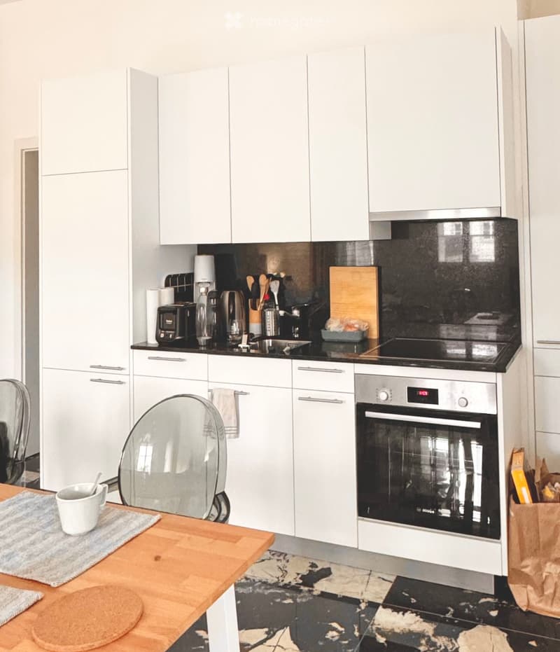 Modern kitchen with white cabinets, black countertops, and stainless steel appliances including an oven, fridge, and coffee maker. A wooden dining table with a chair is visible in the foreground.