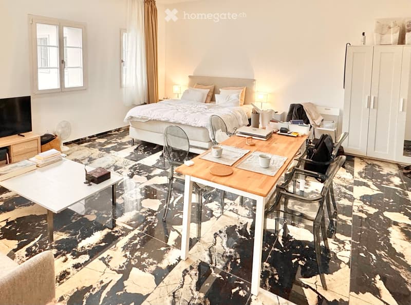 A well-furnished room with a bed, a wooden table with chairs, a laptop, and a fan. The room features large windows with curtains and a patterned rug on the floor.