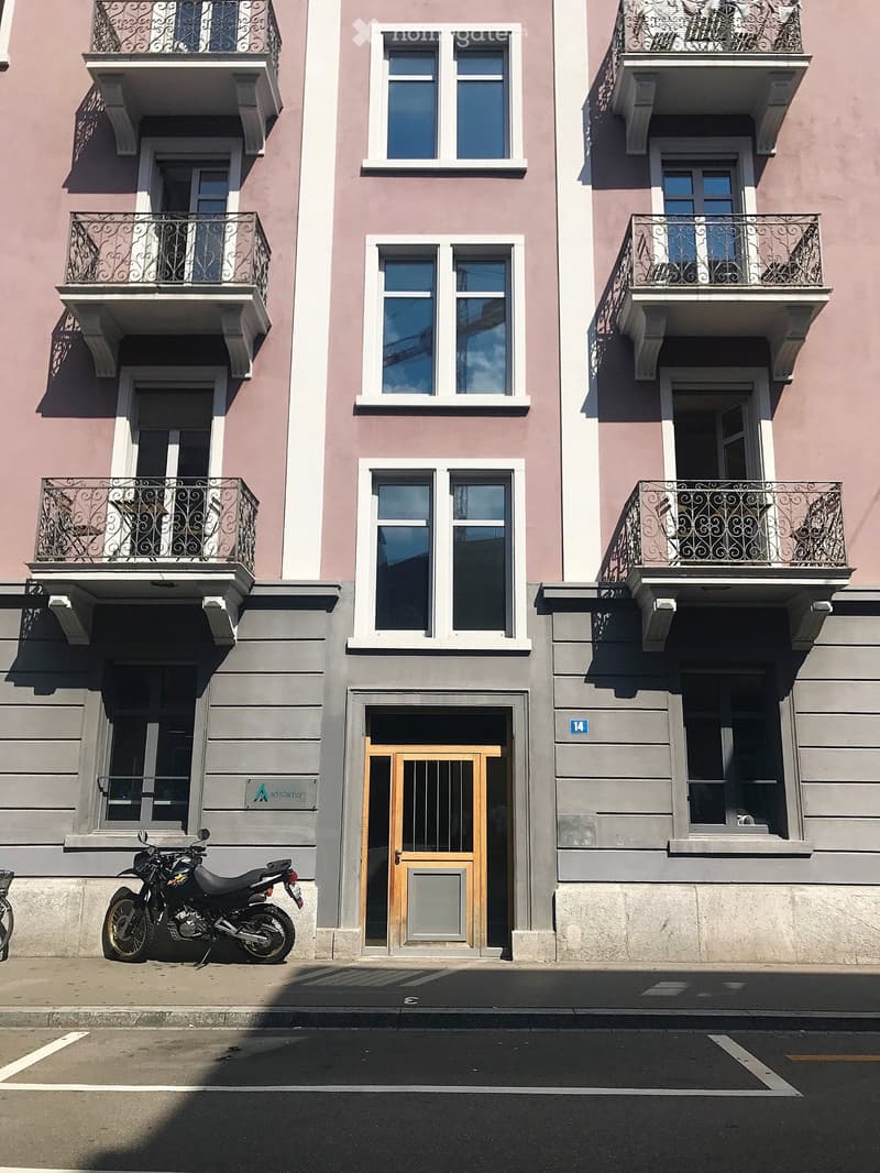 Multi-story building with pink and grey facade, multiple windows, balconies with ornate railings, and a wooden entrance door.