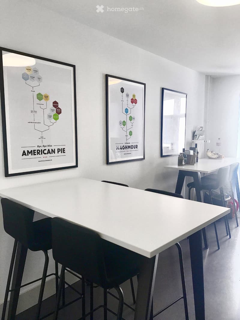 A modern, minimalist room with a large white table surrounded by black chairs and stools, framed posters on the wall, a mirror, and a small counter with a bottle and other items.