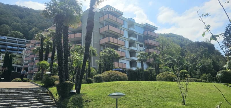 Modern multi-story building with balconies, surrounded by lush greenery and palm trees, with a well-maintained lawn and pathways.
