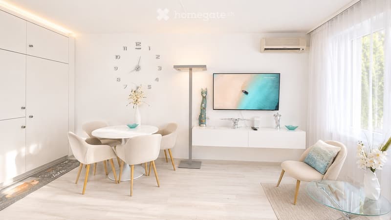 Modern living room with white walls, light wood flooring, and large windows. Features a round dining table with four chairs, a glass coffee table, a sofa with a decorative pillow, a floor lamp, a wall-mounted TV, and a white cabinet with decorative items.