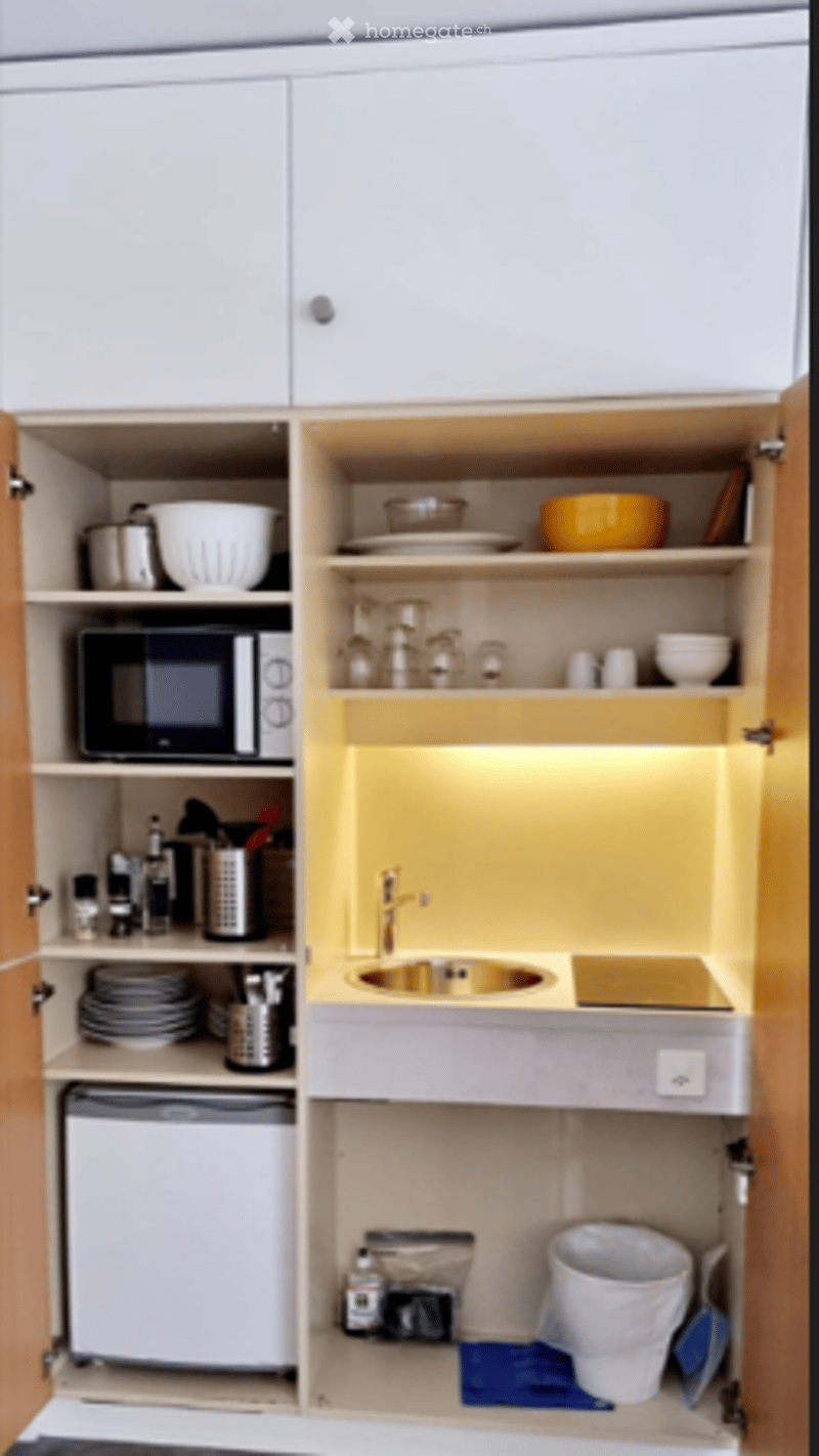 Compact kitchen with white cabinets, microwave, sink, and small refrigerator. Includes dishes, cups, and a toilet in the corner.