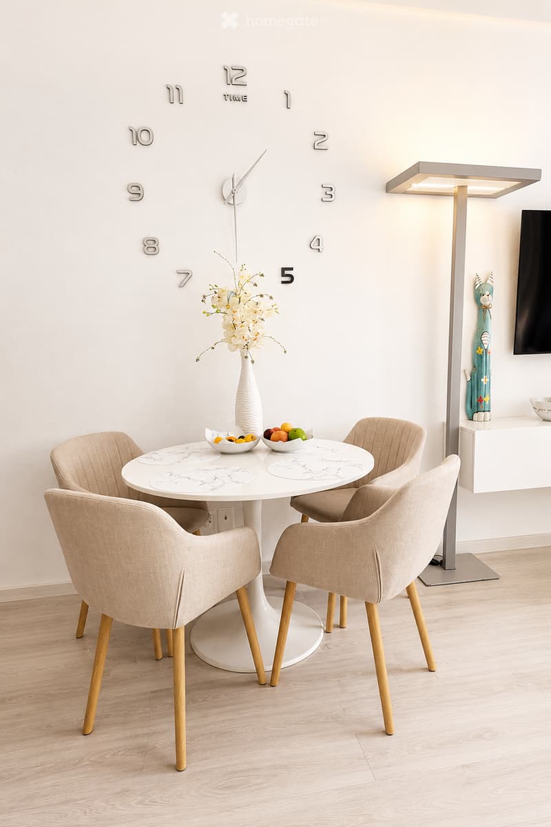 Modern dining area with a white round table, four beige chairs, a vase with white flowers, a decorative clock, a floor lamp, and a screen on a white shelf.