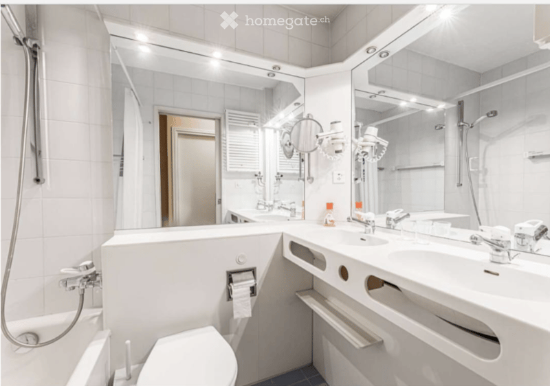 Modern bathroom with white tiles, a toilet, a sink with a mirror, and a shower area. The sink has a modern faucet and a hairdryer is mounted on the wall. The mirror is illuminated with LED lights.