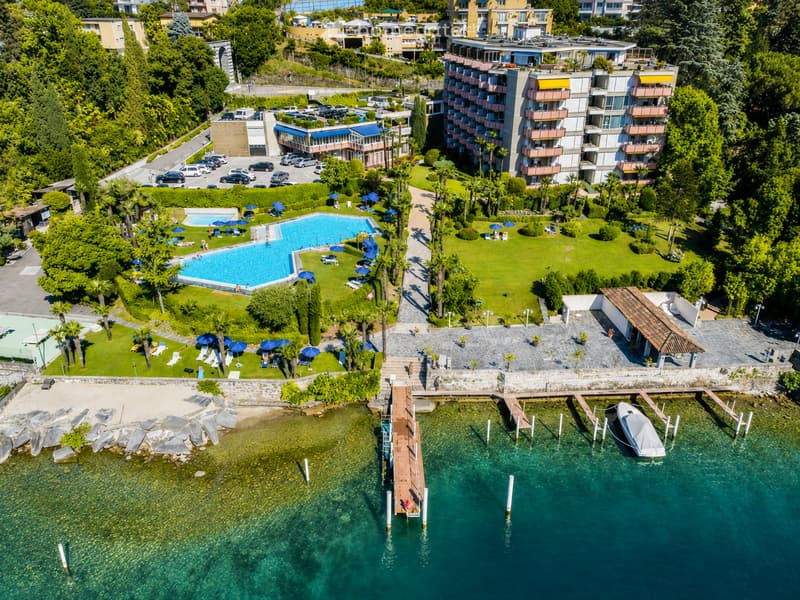 Aerial view of a resort with a swimming pool, lounge chairs, umbrellas, a dock, and a boat on the water, surrounded by greenery and buildings.