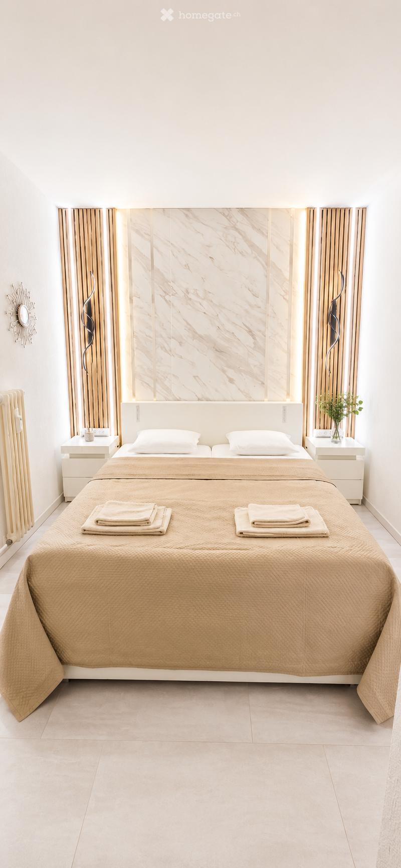 Modern bedroom with a large bed, white nightstands, a decorative mirror, and a radiator. The bed is neatly made with beige bedding and towels placed on it.
