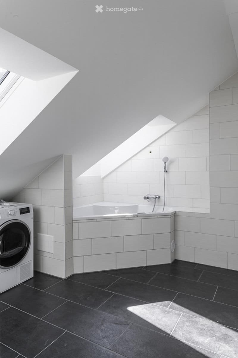 A laundry area with a white washer, tiled walls, a bathtub, and a showerhead. The ceiling features skylights.