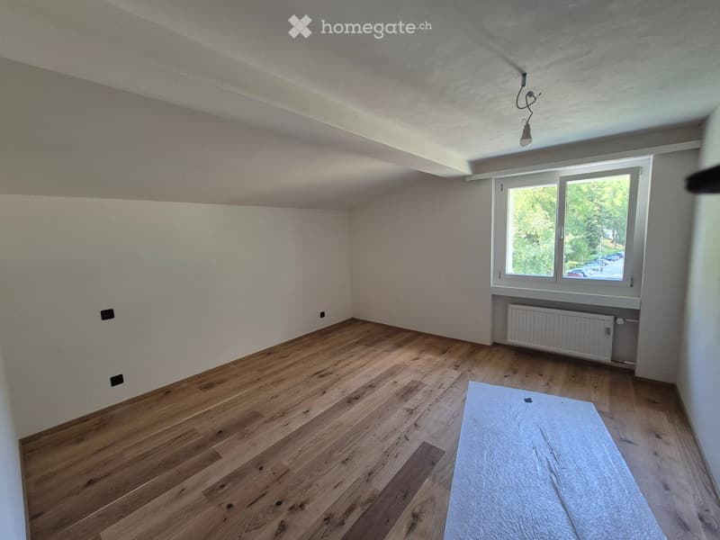 Empty room with wooden flooring, white walls, and a window showing greenery outside. A radiator is visible under the window, and a single light fixture hangs from the ceiling.