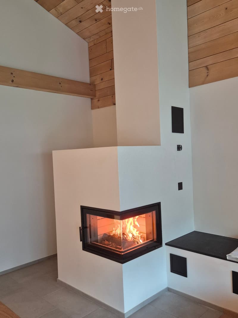 Modern fireplace with a sleek white surround and a black frame around the glass front, showing flames inside. The room has light-colored walls and a wooden ceiling.