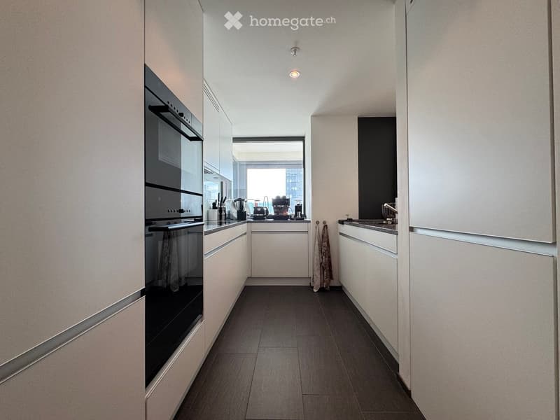 Modern kitchen with white cabinets, dark countertops, and stainless steel appliances including an oven, blender, and coffee maker. A large window provides natural light.