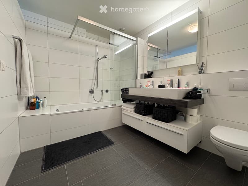 Modern bathroom with a bathtub, sink, toilet, large mirror, and a glass-enclosed shower. Features a dark tiled floor, white tiled walls, and a towel hanging on the side.