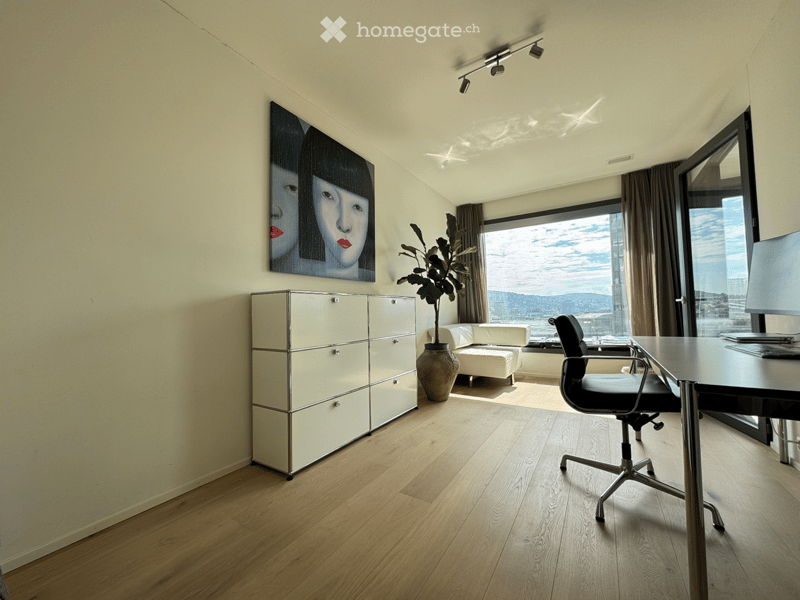 Modern furnished room with wooden floor, large window offering a city view, sofa, desk with chair, cabinet, plant, and artwork on the wall.
