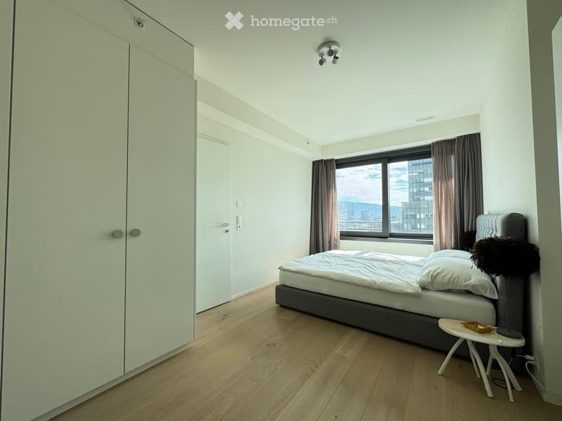 Modern bedroom with a single bed, nightstand, small stool, and large window with curtains. The room features light-colored wooden flooring and white walls.