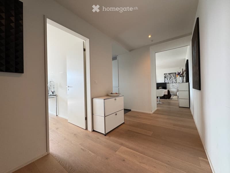 Bright hallway with wooden flooring, white walls, and a small white cabinet. An open door leads to another room with visible furniture.