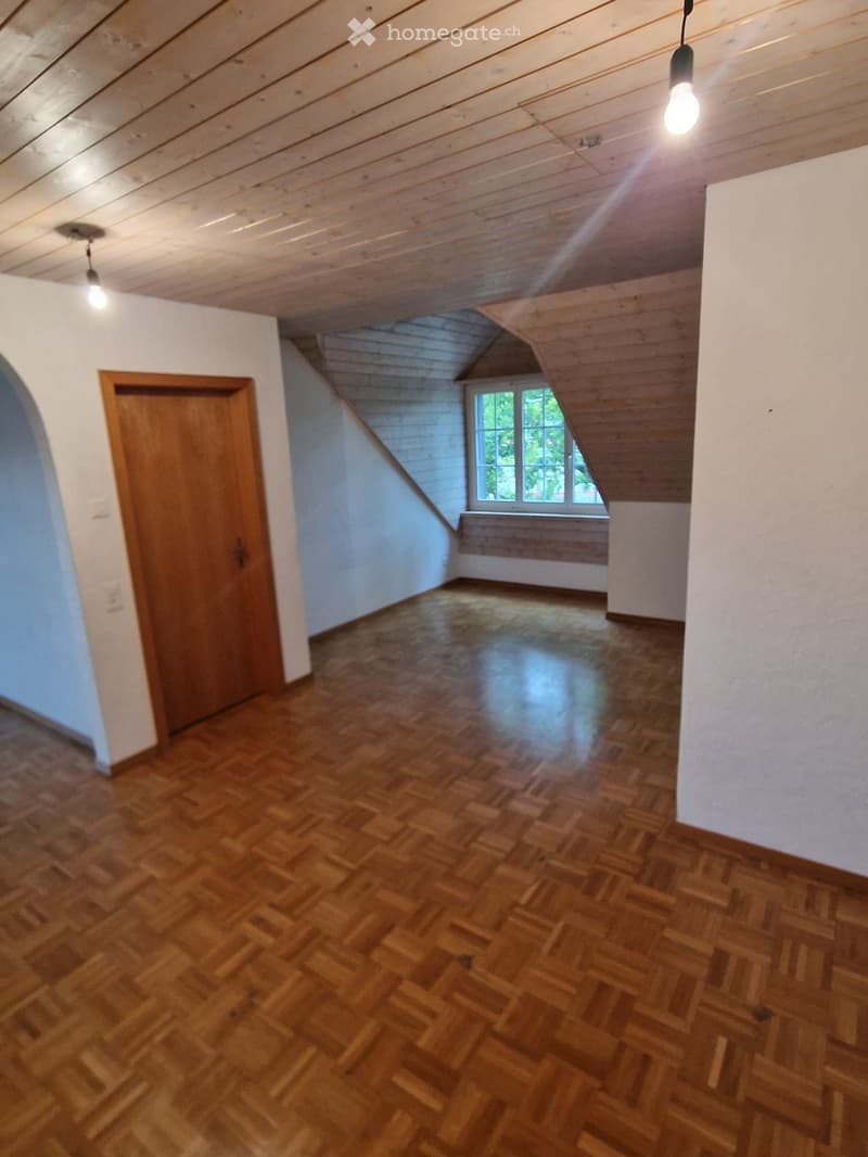 Empty room with wooden floor, white walls, wooden door, and exposed wooden ceiling beams. Natural light from a window on the far wall.