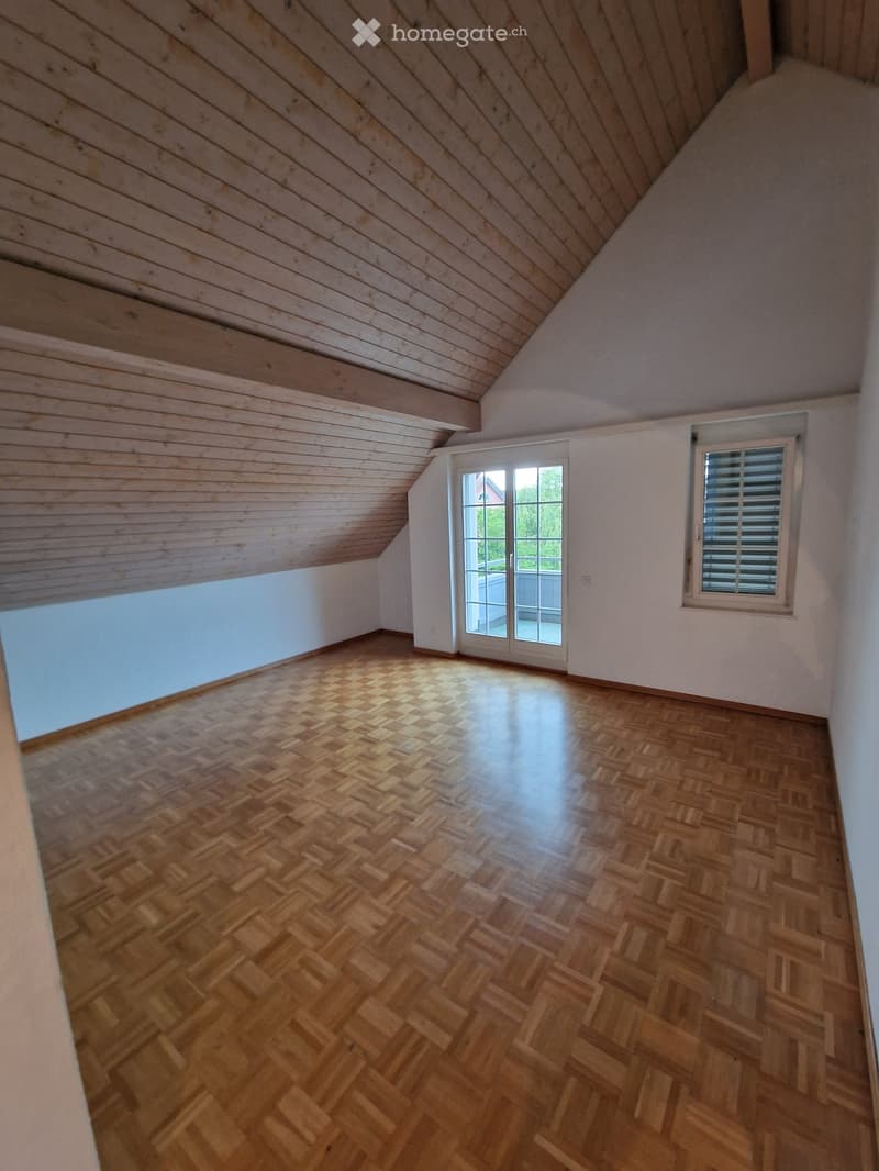 Empty attic room with wooden herringbone flooring, slanted wooden ceiling, white walls, and two windows (one with blinds).