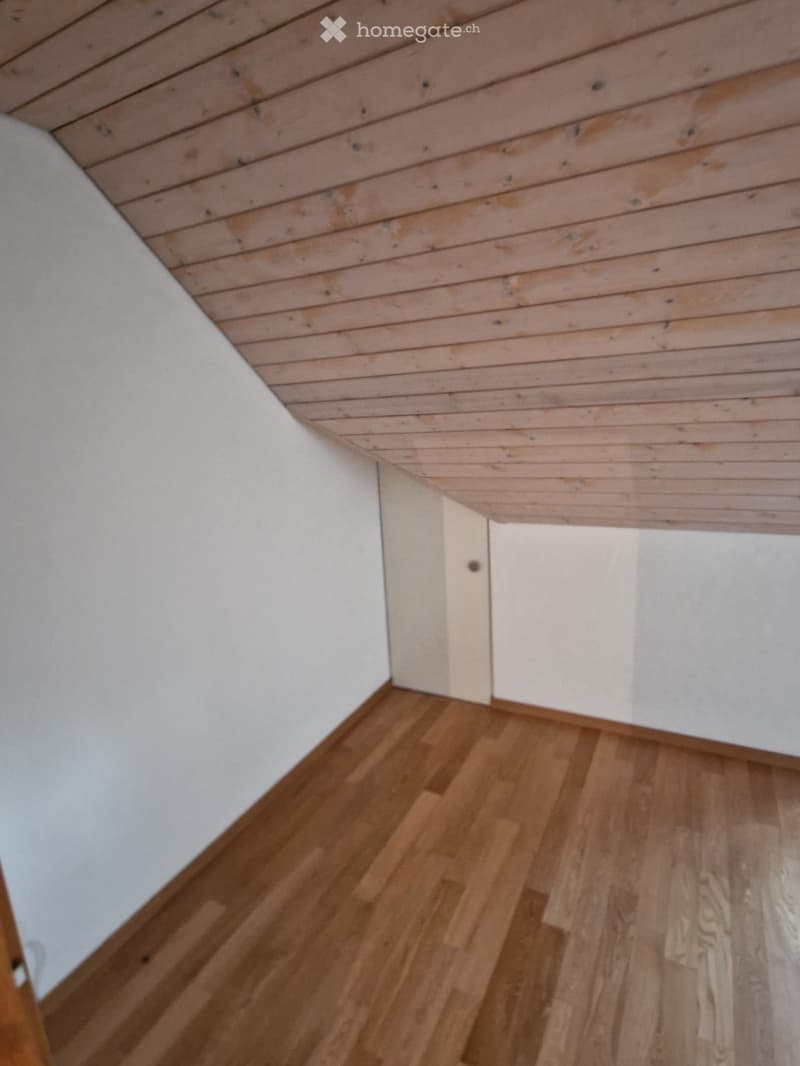 Empty room with wooden flooring and white walls, featuring a slanted wooden ceiling and a small doorway.