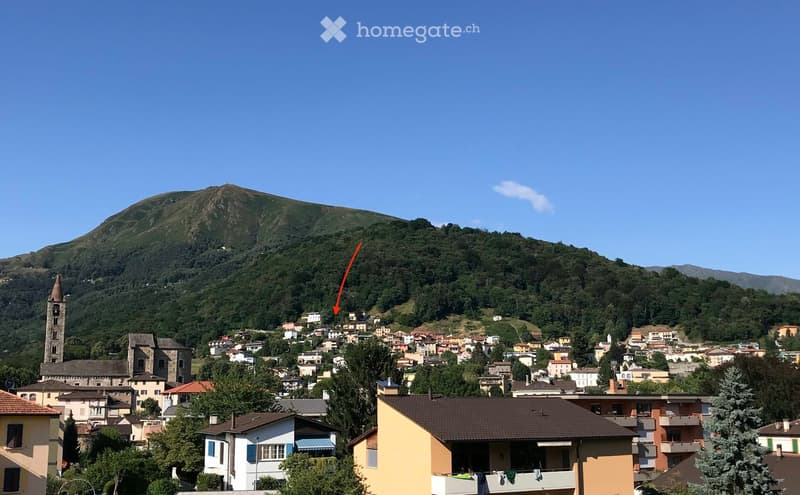 A panoramic view of a small town nestled in a valley surrounded by lush green hills and mountains. The town features a mix of residential and historical buildings, including a prominent church tower, and is surrounded by dense forests and rolling hills.