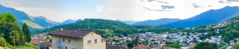 Single-story house with a tiled roof and multiple windows, situated in a scenic valley with mountains in the background and a town below.