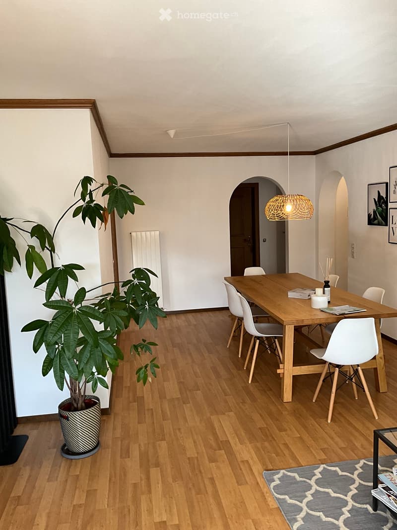 Dining area with wooden table, white chairs, large potted plant, grey rug, and picture frames on the wall.