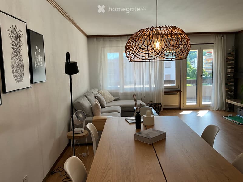 Bright living room with a grey sofa, wooden dining table, chairs, a floor lamp, and a hanging pendant light. Large windows with sheer curtains let in natural light, and a balcony is visible through the glass door.