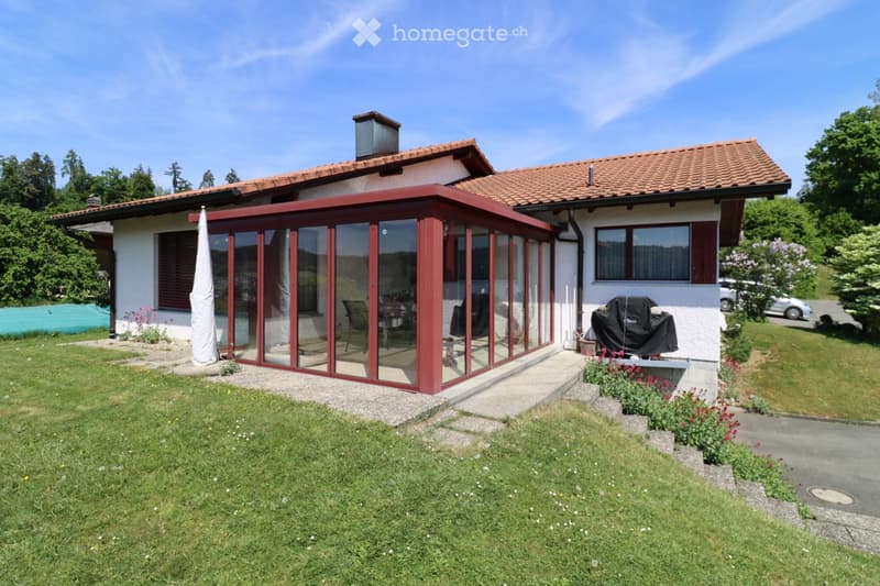 Two-story house with red-tiled roof, white walls, and a large glass-enclosed patio area. The patio has a few chairs and a table, and the surrounding yard is well-maintained with grass and flower beds.