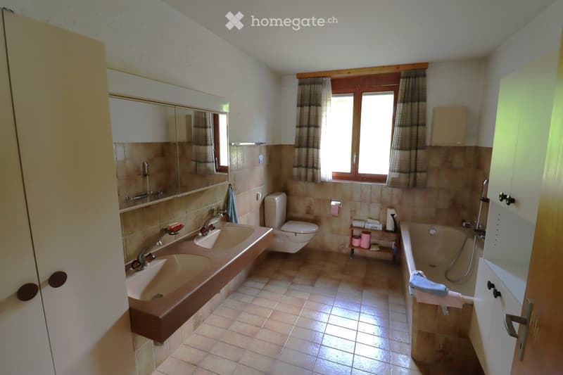 Bathroom with double sink, toilet, bathtub, mirror, and cabinets. Light brown tiles on walls and floor, window with curtains, and a small shelf with towels and toiletries.