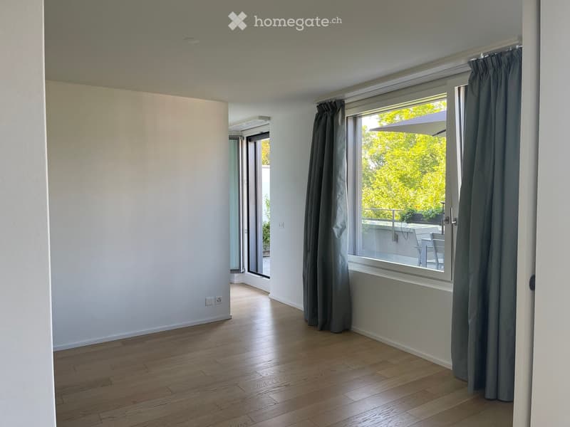 Empty room with wooden flooring, white walls, large window with grey curtains, and sliding glass door leading to a balcony.
